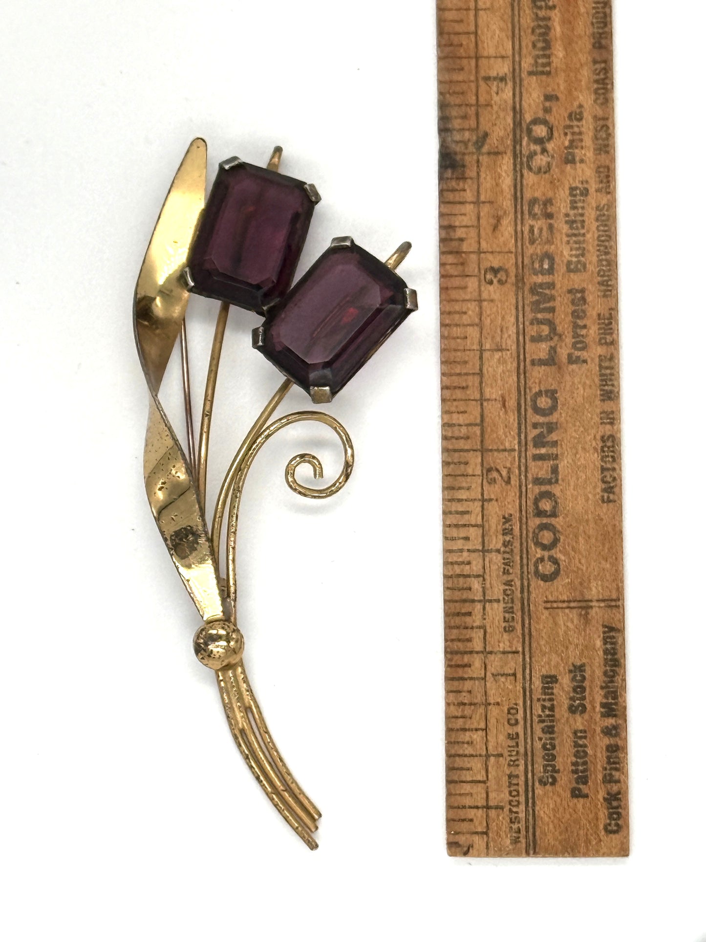 A vintage gold-tone brooch with two rectangular dark purple cabochon gemstones, shown next to a wooden ruler for scale on a white background.