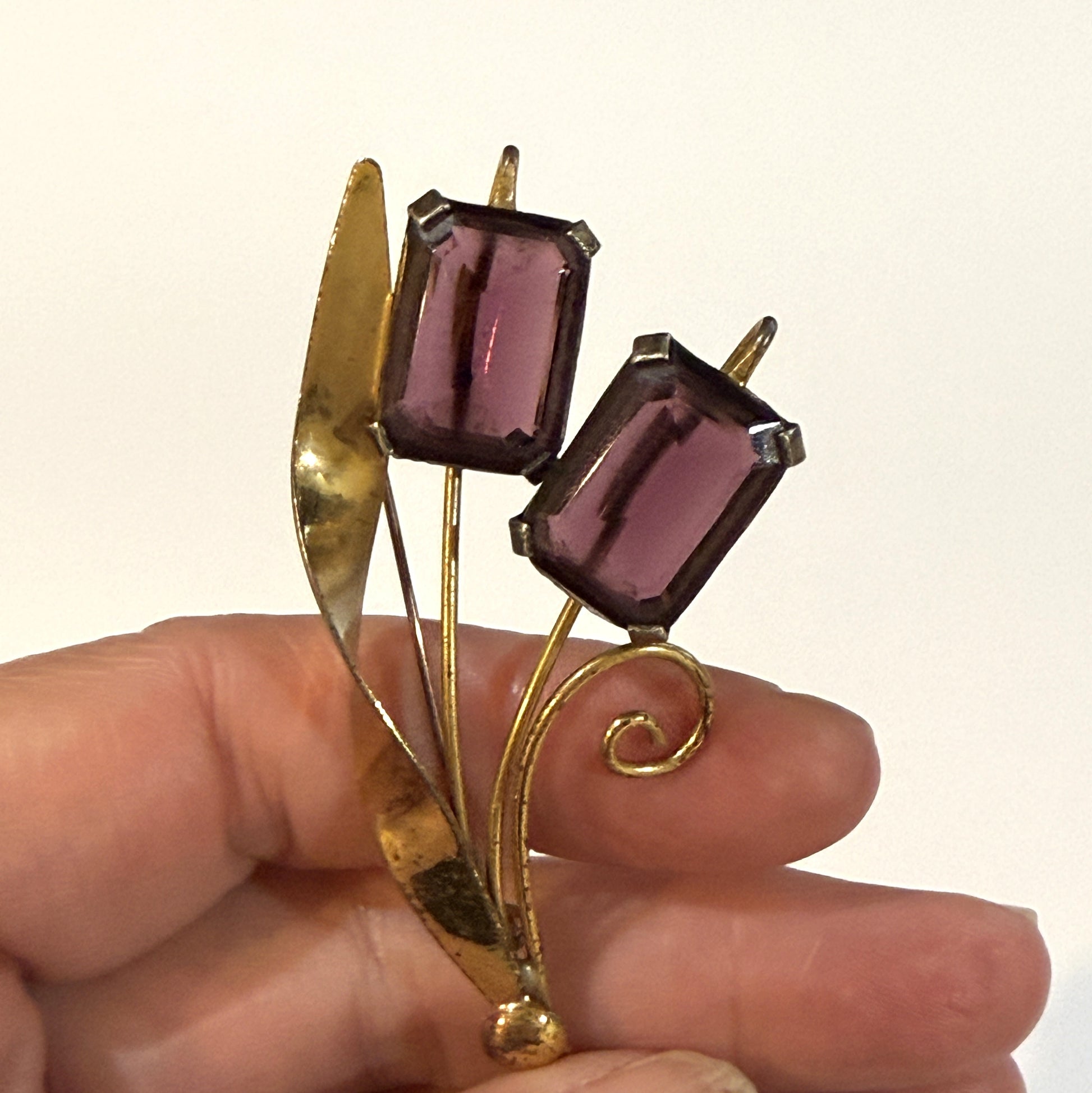 A vintage gold-tone brooch featuring two rectangular purple glass flowers on a leaf-shaped stem, held in a person's hand against a plain light background.